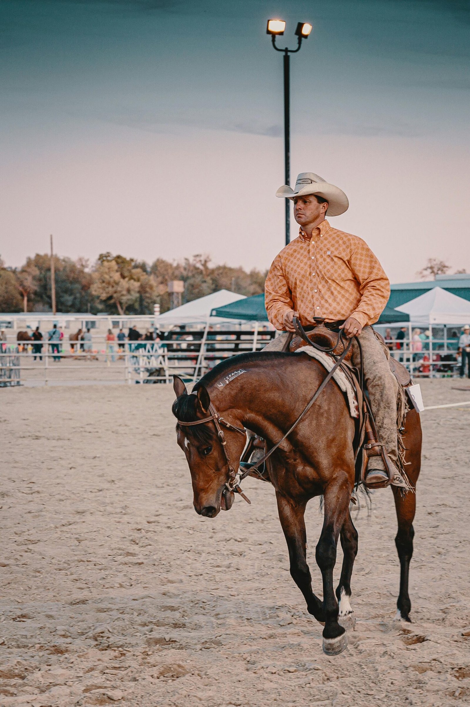 A vertical shot of a man in a tan western shirt and cowboy hat riding a brown horse in an